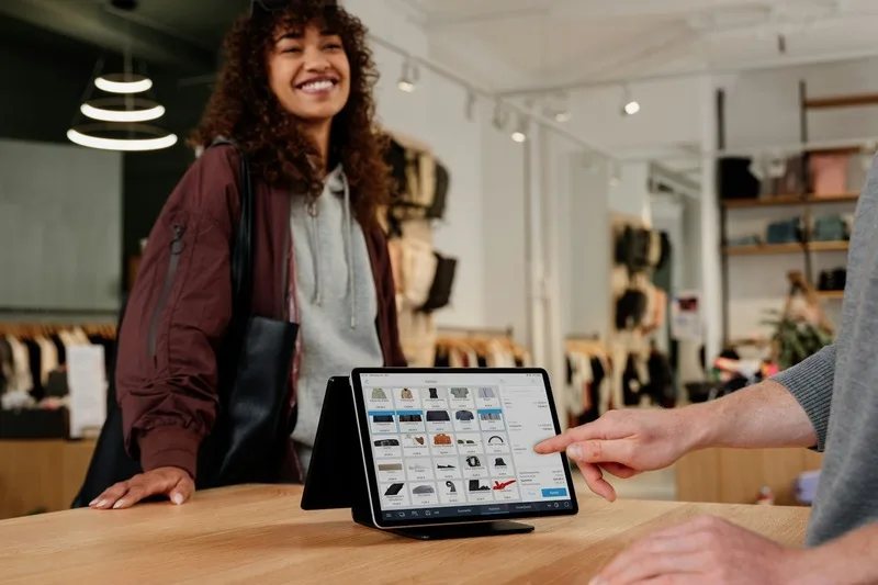 A retail associate using a tablet point-of-sale system on a store counter while helping a customer, representing omnichannel retail for Shopify Plus