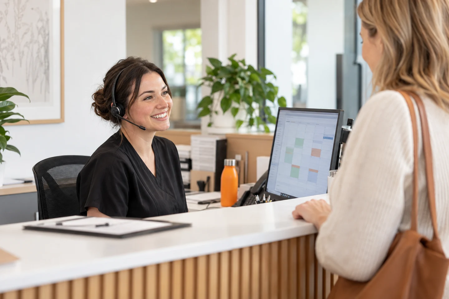 AI-generated photo showing A clinic receptionist on a headset greeting a patient at the front desk, with a booking calendar on her monitor