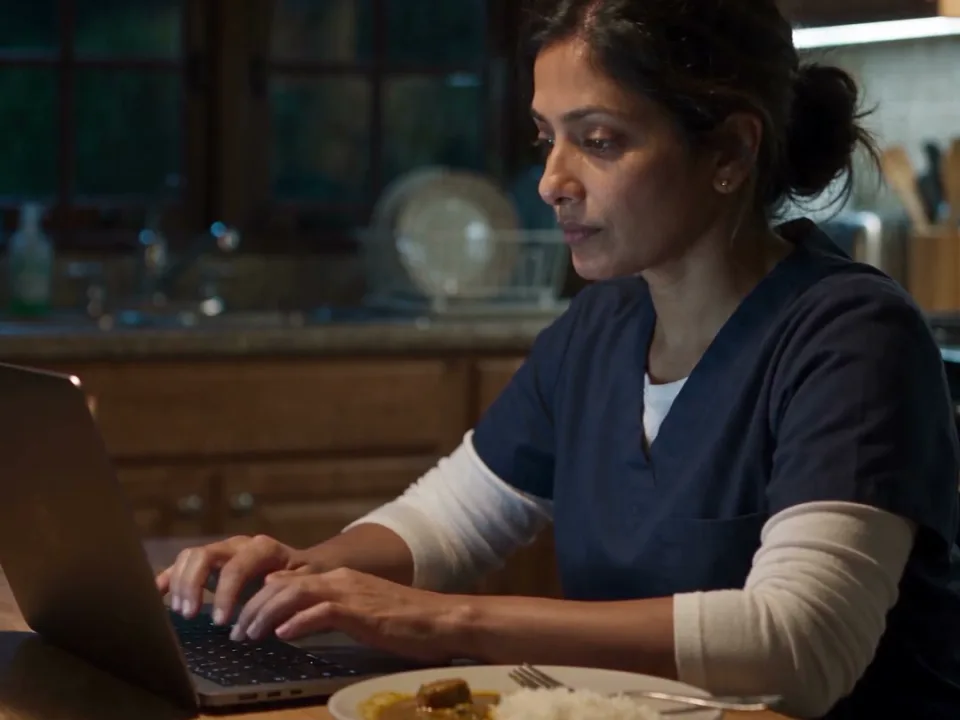 AI-generated still from our film: a doctor in scrubs types notes on a laptop at her kitchen table at night, dinner beside her.